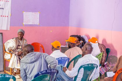 A group of people, some wearing Ipas orange caps, sits in colorful plastic chairs. They are listening attentively to a person dressed in traditional attire seated in front of a banner about gender-based violence and a chalkboard. The room has purple and pink walls.