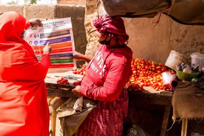 An individual in bright orange holds up an informational poster to show a woman who is wearing red as they stand outside.